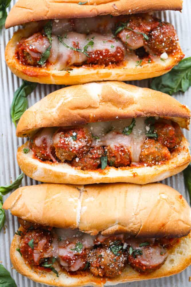 Three meatball sub sandwiches laid out on a metal sheet pan.  Melted cheese on top of meatballs in toasted hoagie rolls.  The sub is decorated with fresh basil leaves on the side.