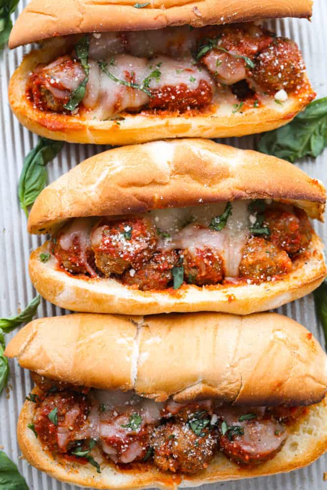 Three meatball sub sandwiches laying on a metal sheet pan. Melted cheese on top of the meatballs in a toasted hoagie roll. The subs are garnished with fresh basil leaves on the side.