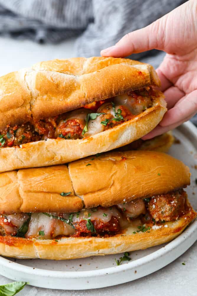 Close up view of a stack of meatball subs.  A hand is reaching in and lifting up one of the subs on top.