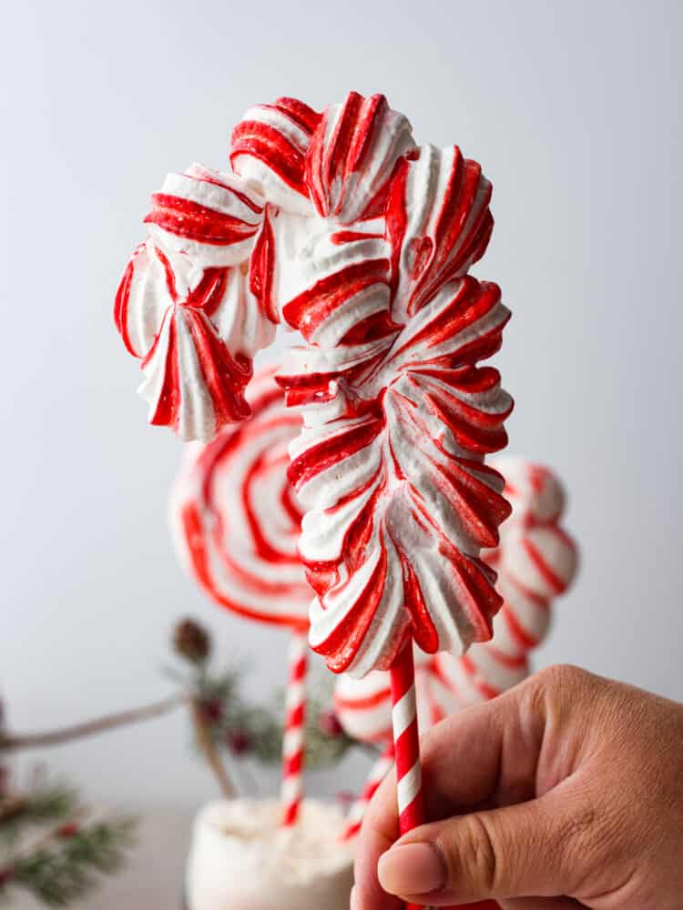 Peppermint meringue lollipops being held by a hand held up by a striped red and white straw.