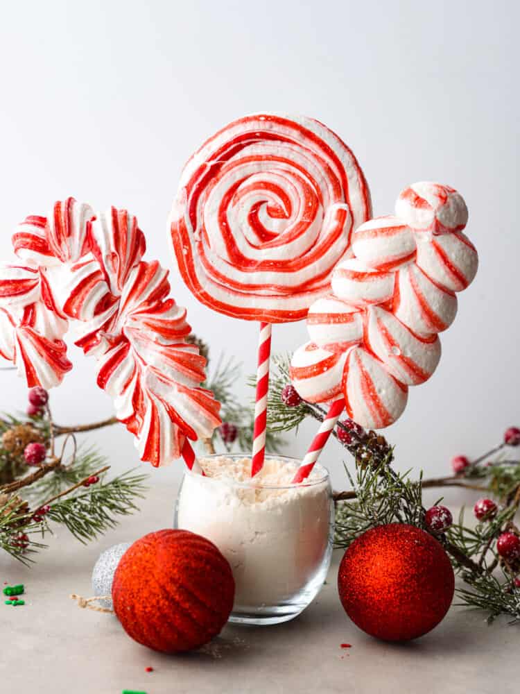 Peppermint meringue lollipops in a cup with red ornaments and green branches.