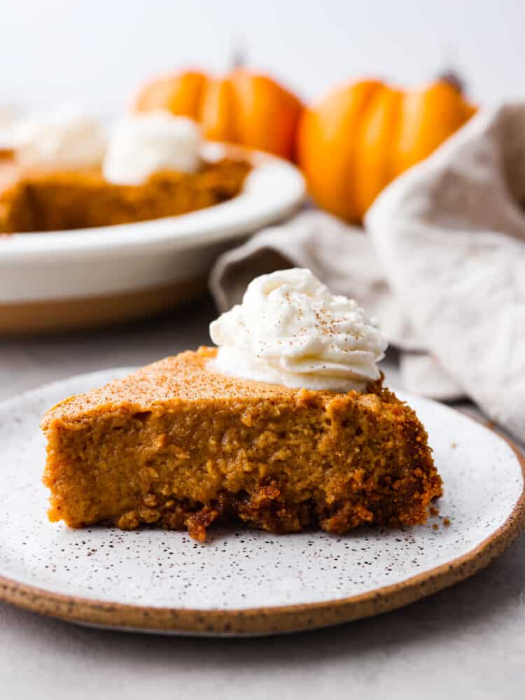A slice of pumpkin chiffon pie sitting on a stone like plate with the full pie in the background along with pumpkins and a tea towel.