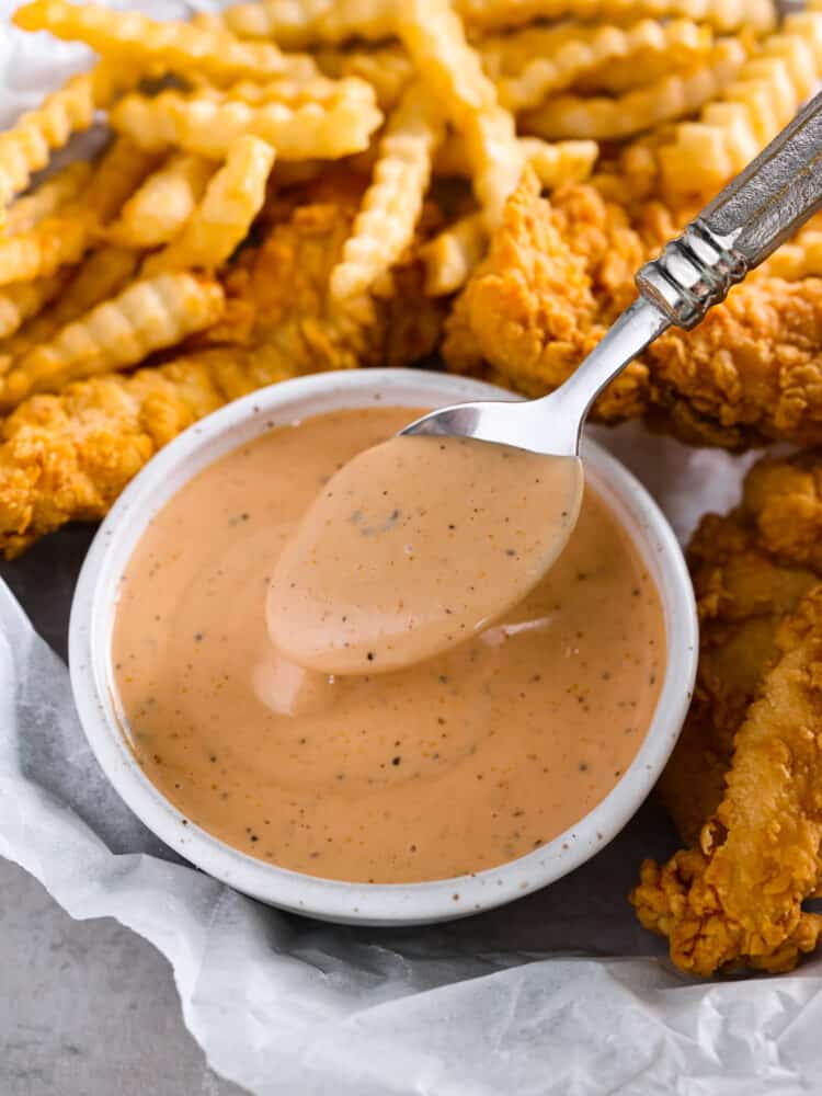 Close up view of sauce in a small white bowl on top of parchment paper. A spoon is lifting up sauce. French fries and chicken stripes are scattered around the bowl.