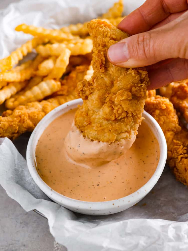 Close up view of a chicken finger being dipped into the bowl of sauce. French fries and chicken stripes are scattered around the bowl.