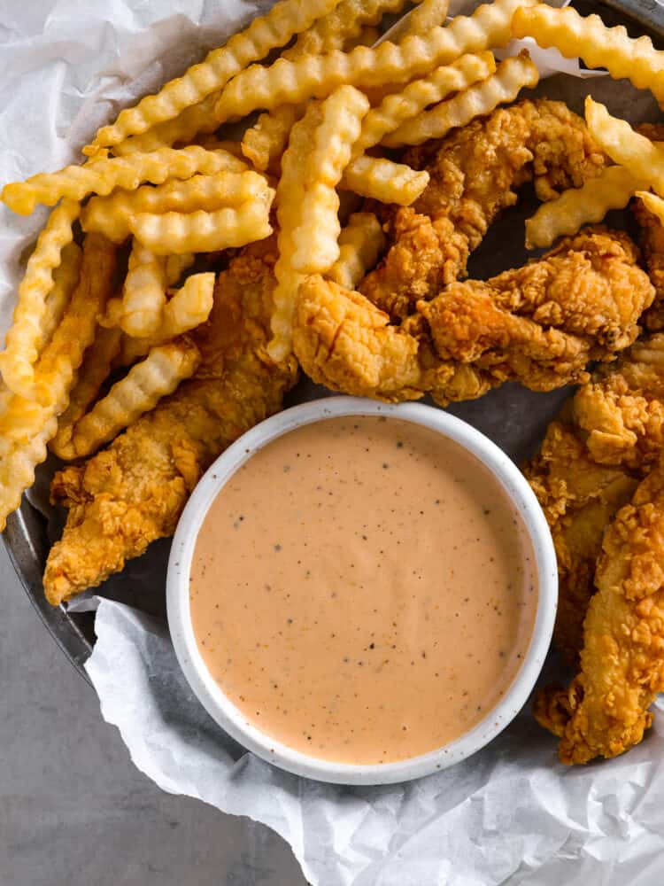 A top view of the sauce in a small white bowl. The bowl is on a round tin plate with parchment paper. Chicken fingers and crinkle cut fries are surrounding the bowl.