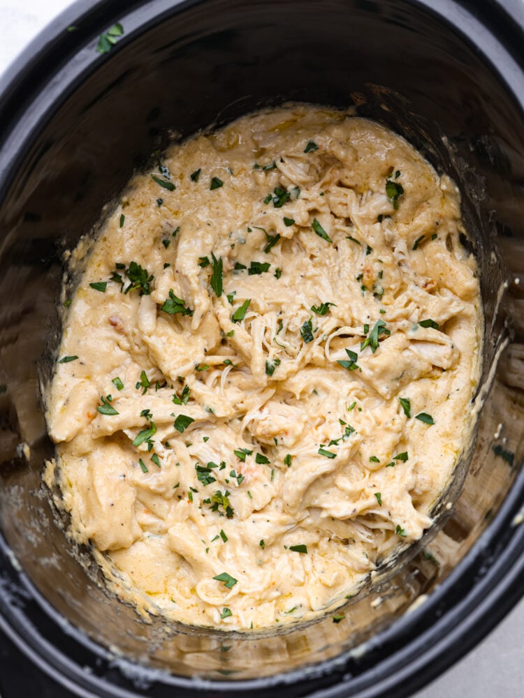 Close up top view of cooked slow cooker angel chicken in a black crockpot.  Fresh parsley is garnished on top.
