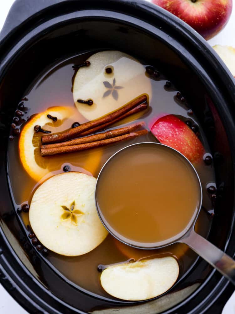 A slow cooker filled with apple cider, a measuring cup, apple slices and cloves. 