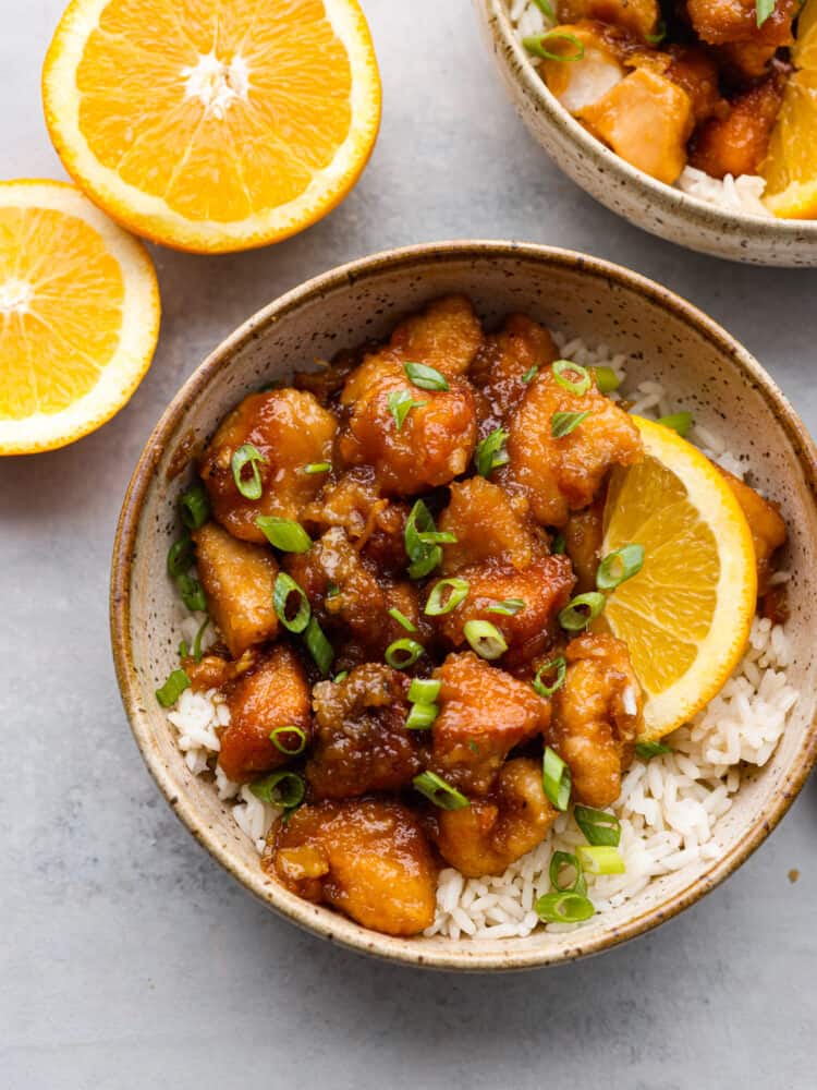 The top view of a bowl of orange chicken on rice with some orange slices. 