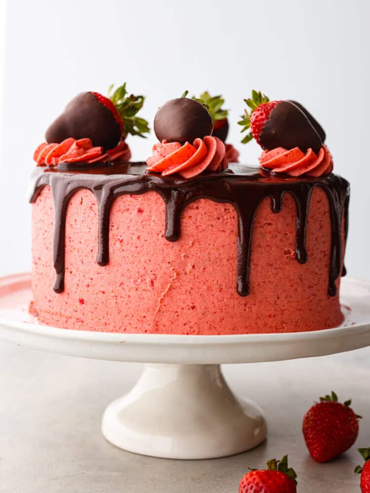 Front view of strawberry chocolate cake on a white cake stand and white background.  Whole strawberries are garnished on the side of the cake stand.