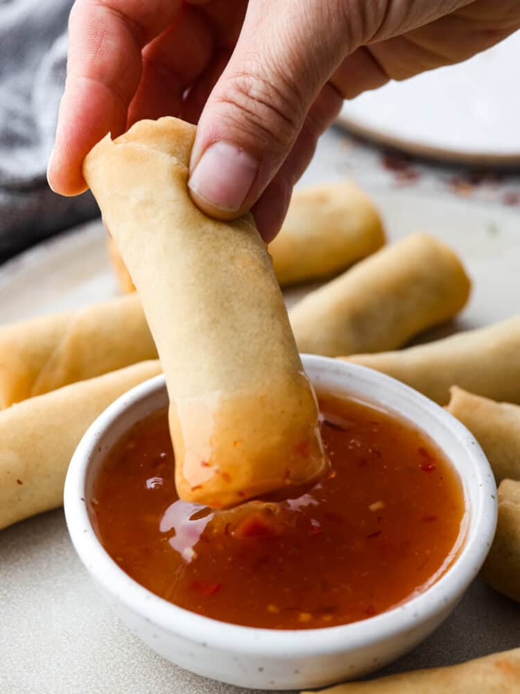 A side view of a small bowl full of sweet chili sauce with an egg roll being dipped into the sauce.  The bowl of sauce is on a gray plate with egg rolls scattered all around the plate close to the sauce.