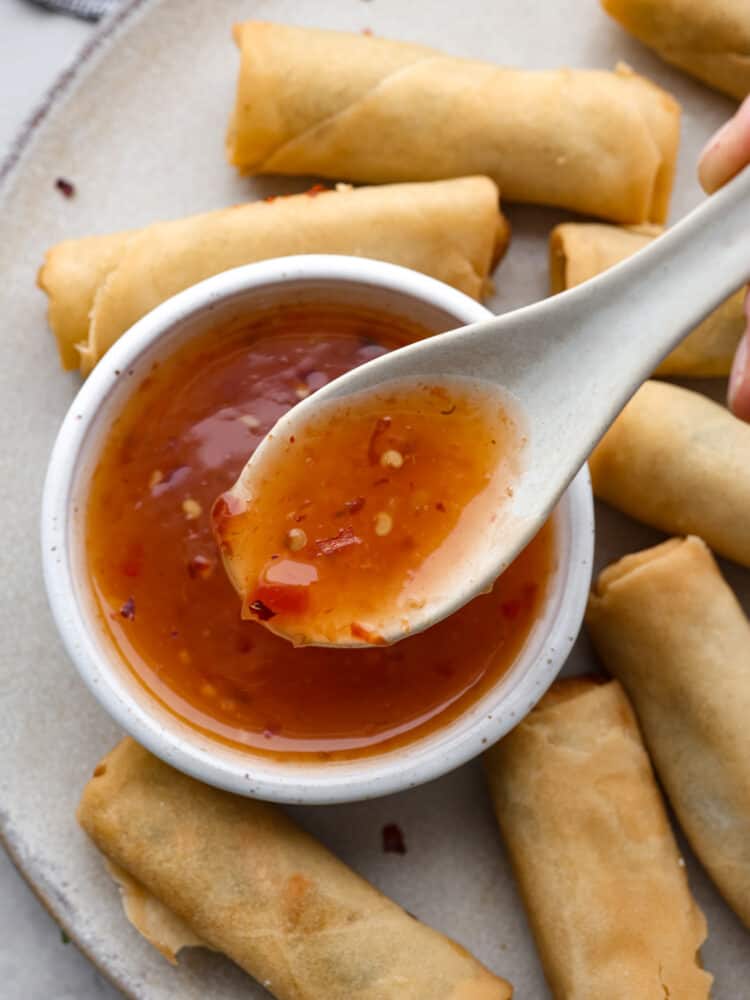 A top view of a small bowl full of sweet chili sauce and a spoon lifting out a spoonful of sauce. The bowl of sauce is on a gray plate with egg rolls scattered all around the plate close to the sauce.