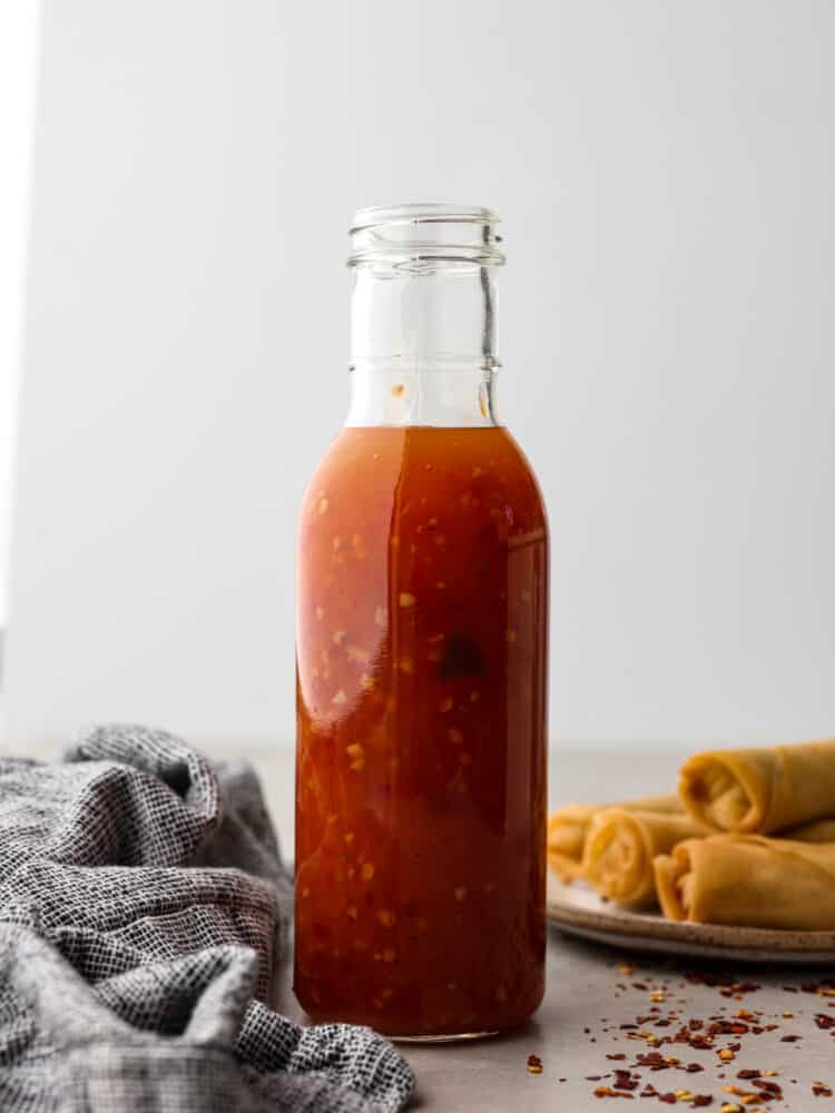 Side view photo of sweet chili sauce in a tall glass bottle with a gray kitchen towel and a plate of egg rolls next to the bottle.  Red chili flakes are scattered on top of the gray countertop.