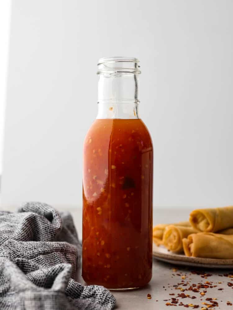 Side view photo of sweet chili sauce in a tall glass snifter with a gray kitchen towel and a plate of egg rolls next to the bottle.  Red chili flakes are scattered on top of the gray countertop.