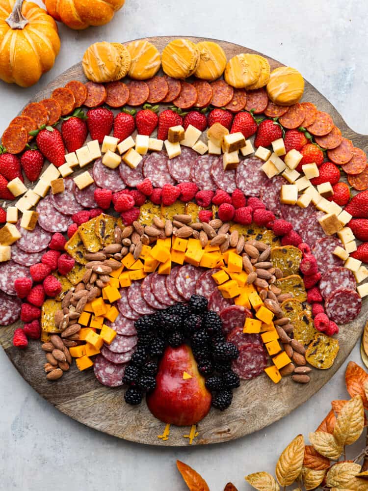 Overhead view of a turkey charcuterie board. Pumpkins and fall leaves are garnished around the wood board.