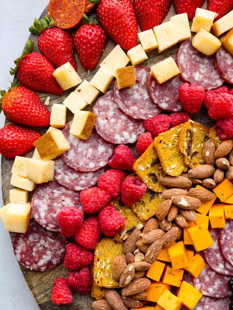 Close overhead view of charcuterie ingredients. Strawberries, salami, cheeses, raspberries, crackers, and almonds are arranged on the board.