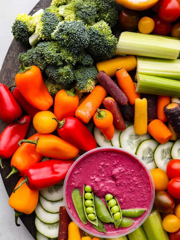 Top view of the beet hummus with sugar snap peas garnished on top. Veggies are surrounding the hummus.