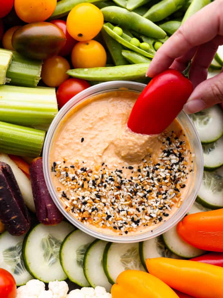 Close up of a red bell pepper dipping into the hummus. Veggies surround the hummus.