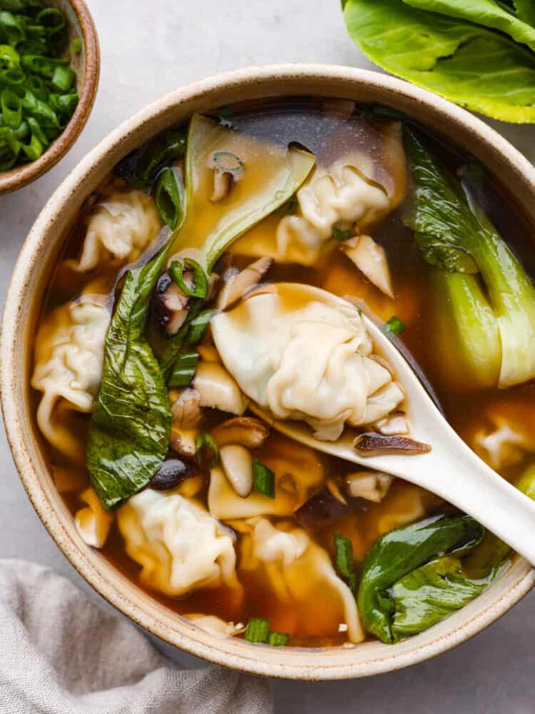 Top view of wonton soup in a tan bowl with a white spoon lifting up a wonton dumpling. A tan towel and bowl of green onions are next to the bowl.