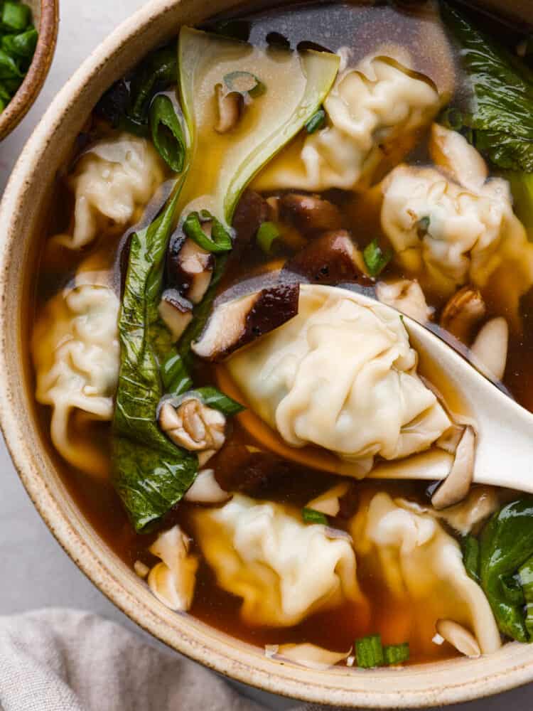 Close up photo of wonton soup in a tan bowl with a white spoon lifting up a wonton dumpling.  A tan towel and bowl of green onions are next to the bowl.