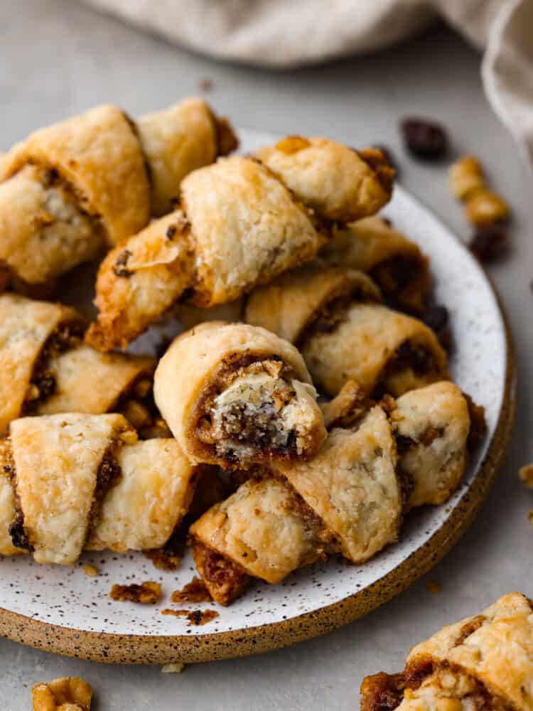 A closeup photo of a small plate of rugelach cookies stacked on top of each other. Raisins, walnuts, and a kitchen towel are scattered around the plate. One of the cookies has a bite taken out of it.