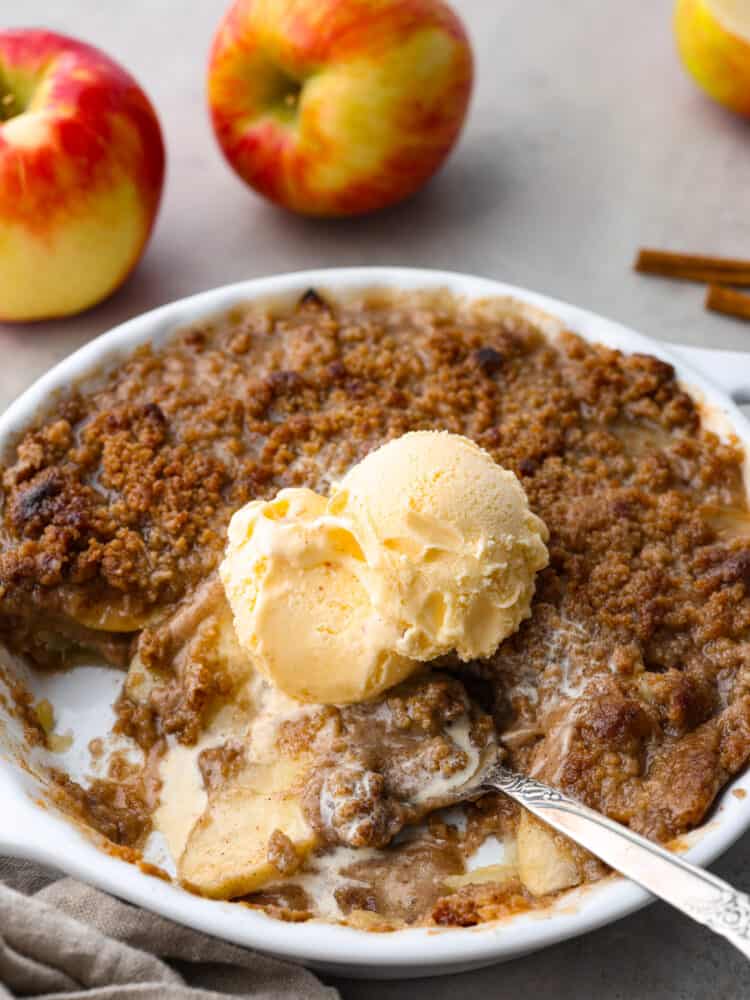 Apple Brown Betty in a white dish with vanilla ice cream on top and scoops taken out. A metal spoon is sitting in the dish with apples in the background