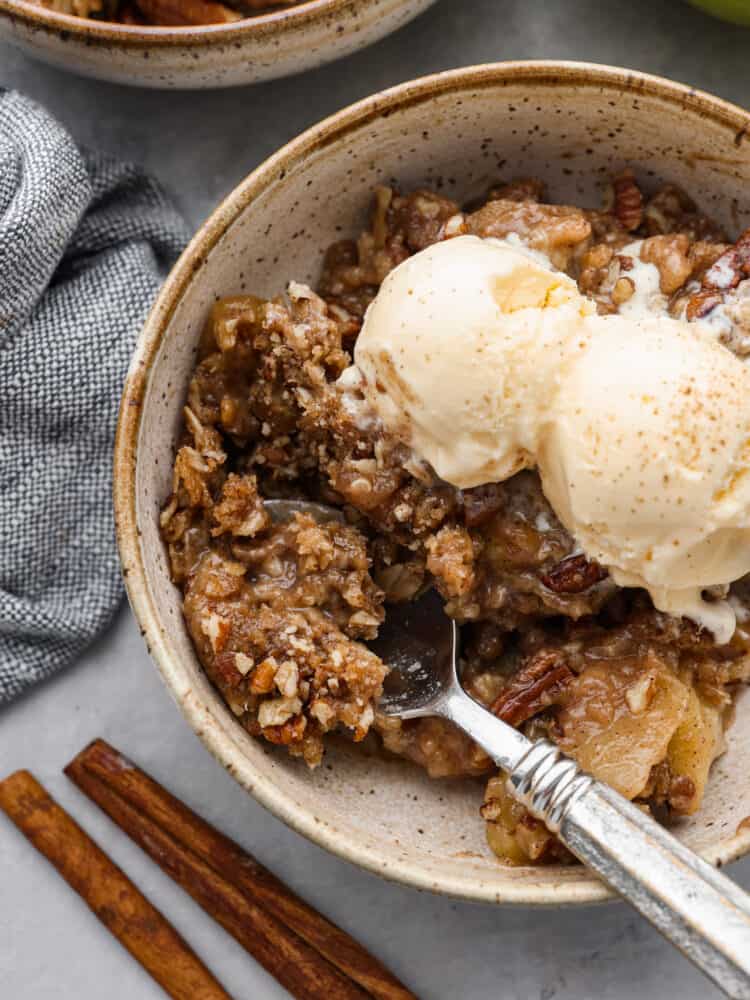A bowl of apple pecan crisp with ice cream in a bowl with a silver spoon scooping some out. 