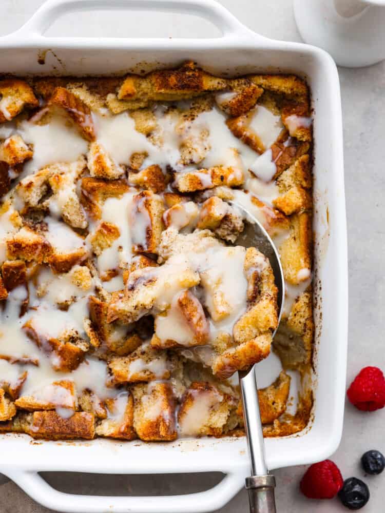 Bread pudding in a white pan with vanilla sauce on top and a metal spoon taking a scoop out.