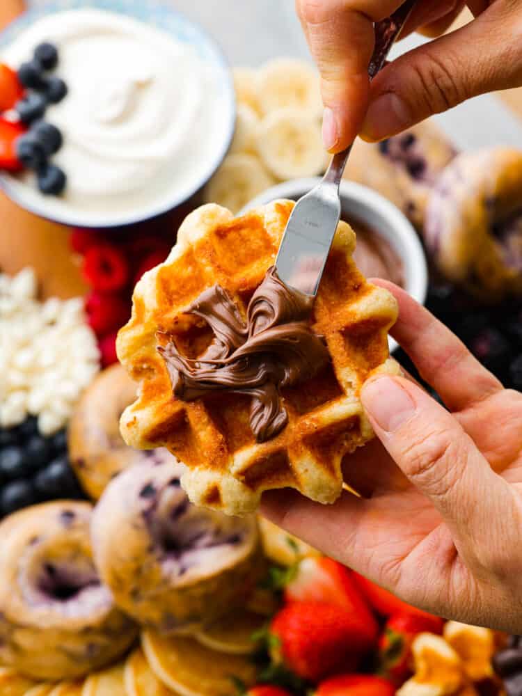 Close up view of Nutella being spread on a waffle with the board of ingredients in the background.