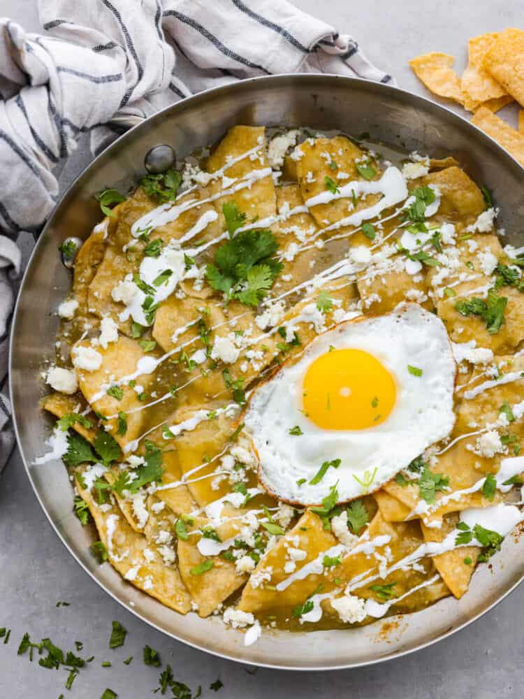 Chilaquiles in a pan with a fried egg on top with a tea towel and tortilla chips in the background.
