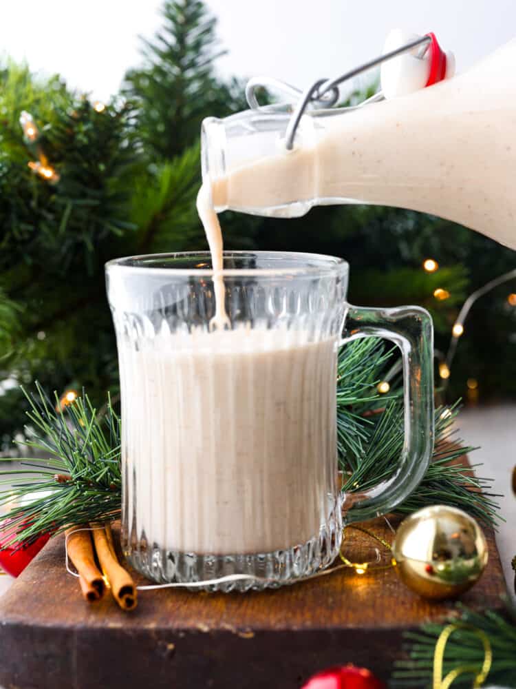 Coquito is poured into a glass with green branches and lights behind it.