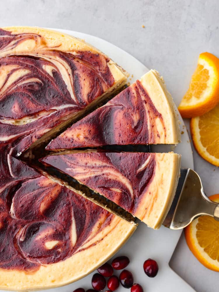 A whole orange cranberry cheesecake with two slices slightly removed on a plate with oranges and cranberries to the side.
