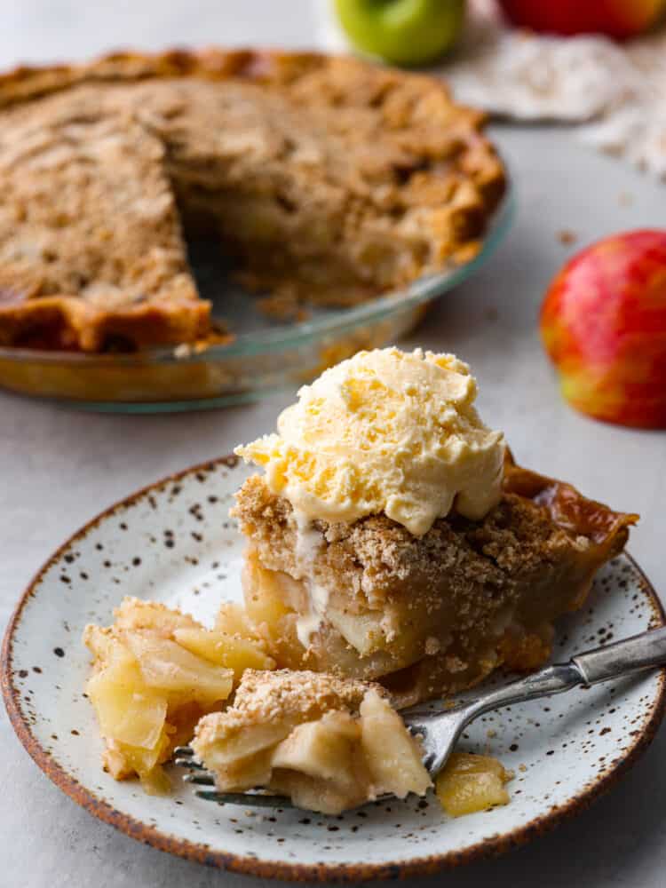 A slice of apple pie served with ice cream on top. There is a whole apple pie in the background.