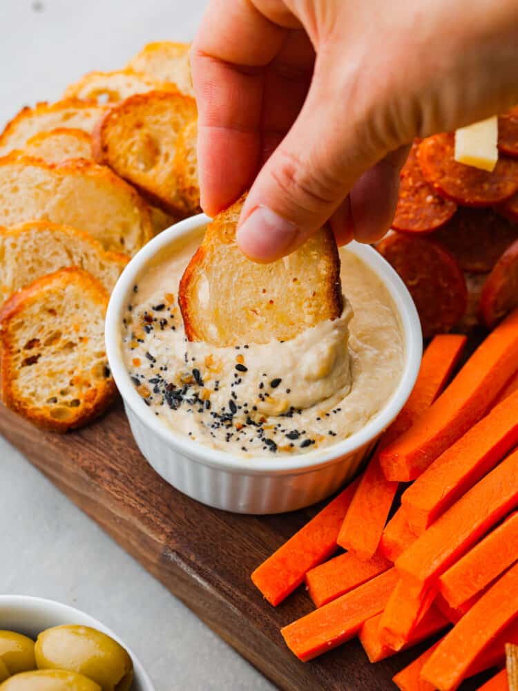 A slice of crostini being dipped in some hummus on the charcuterie board. 