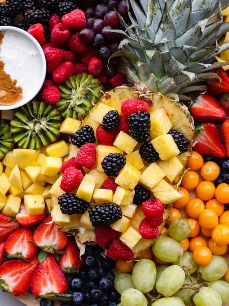 Close overhead view of the sliced whole pineapple filled with pineapple chunks and berries.  The rest of the fruit is scattered around the pineapple with fruit dip on the side.