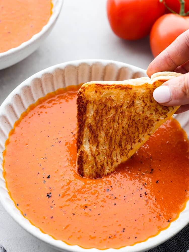 Tomatsoppa i en vit skål med en hand som doppar en bit rostat bröd i den.