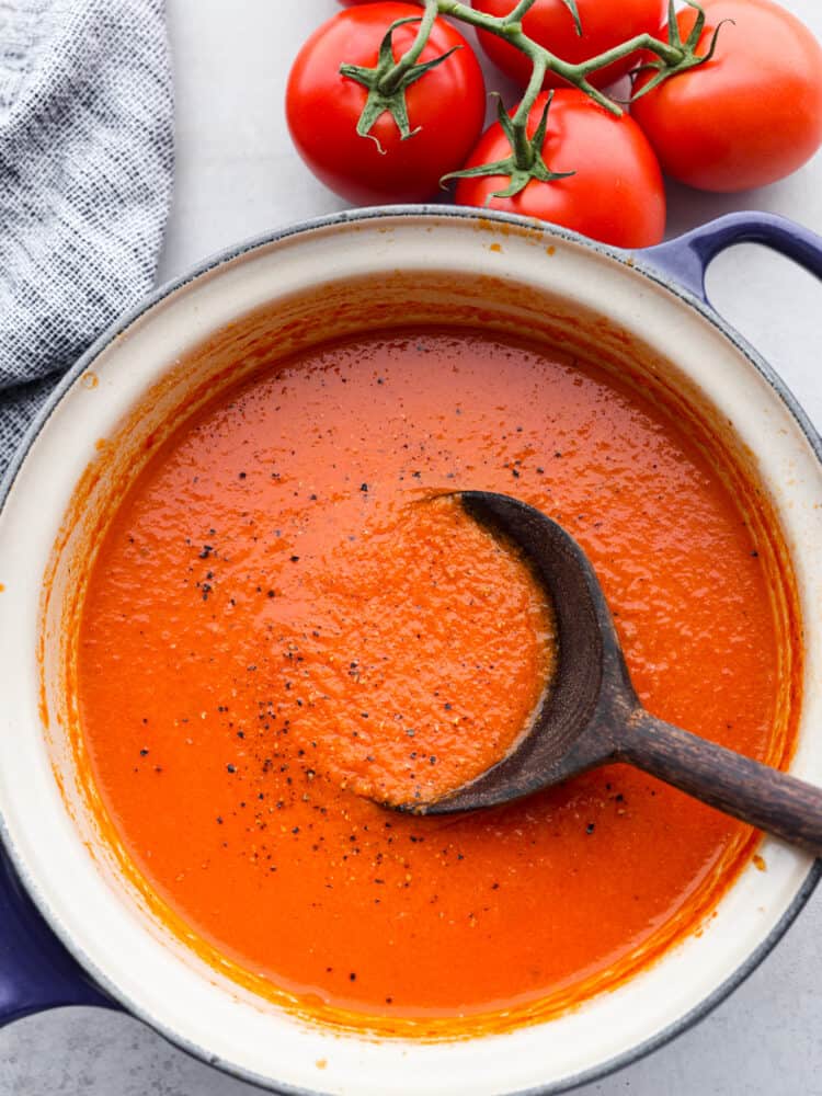 Tomato soup with a black wooden spoon sitting in it with ripe tomatoes and a blue tea towel to the side.