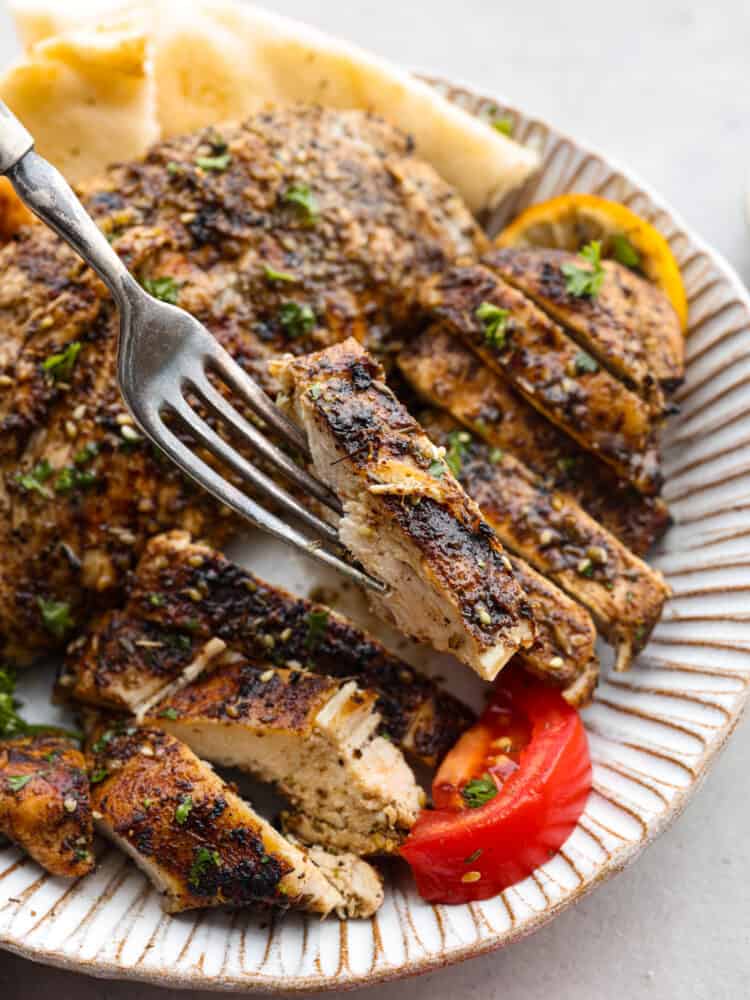 A fork holding up a piece of chicken over the plate of Za'atar chicken with a tomato and lemon.