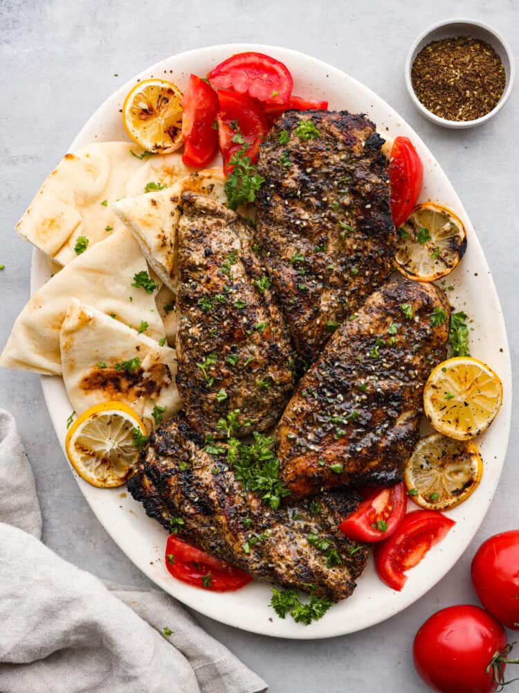 Za'atar chicken on a plate with tomatoes, lemons, pita bread, and seasoning to the side.