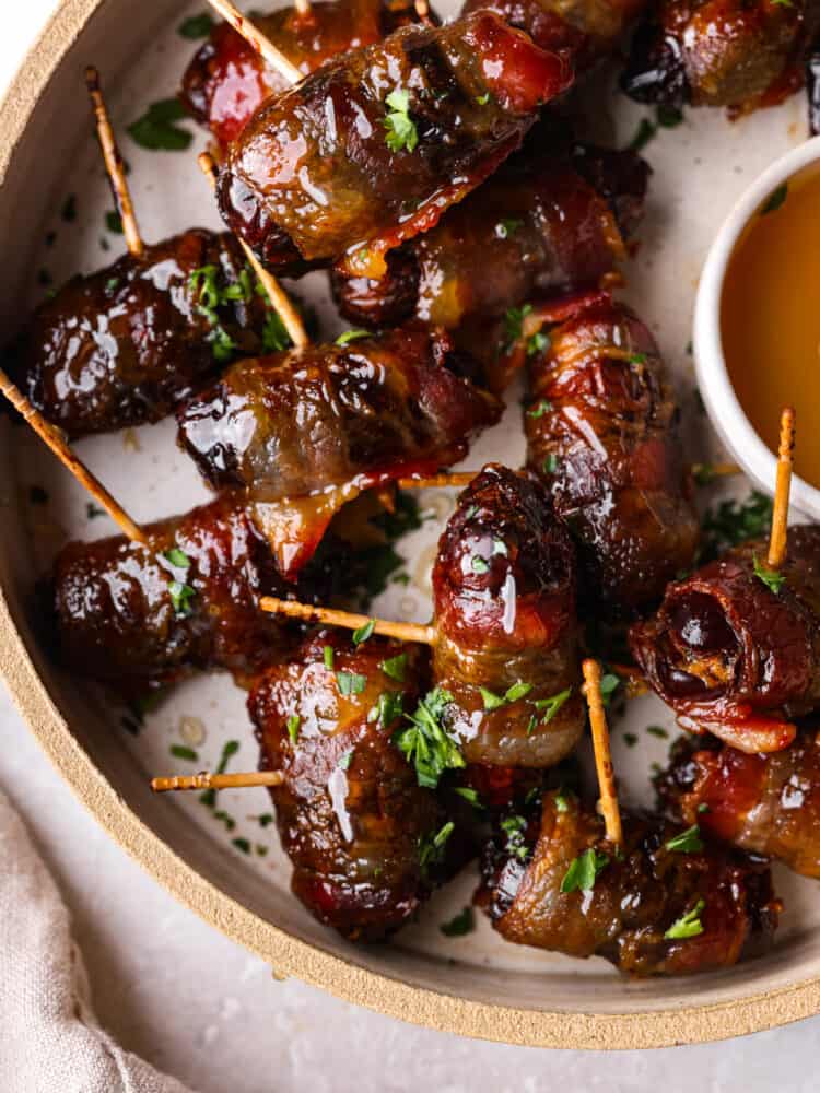 Close up view of bacon wrapped dates on a tan colored plate.  A small bowl of honey is on the plate with a tan colored kitchen towel styled next to the plate. Chopped parsley is sprinkled on top.