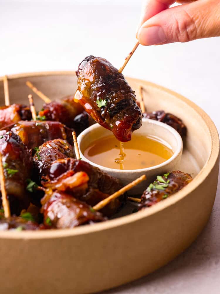 A close up side view of a bacon wrapped date being dipped into a small bowl of honey.