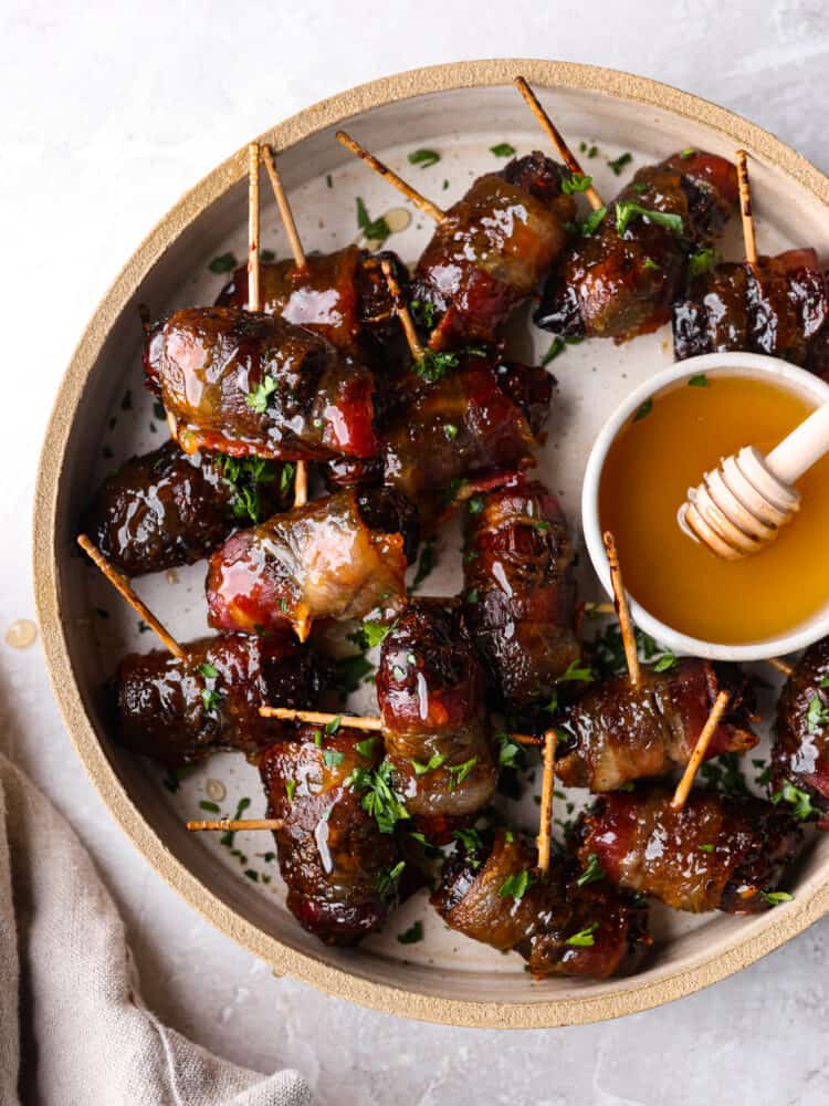 Top view of bacon wrapped dates on a tan colored plate. A small bowl of honey is on the plate with a tan colored kitchen towel styled next to the plate.