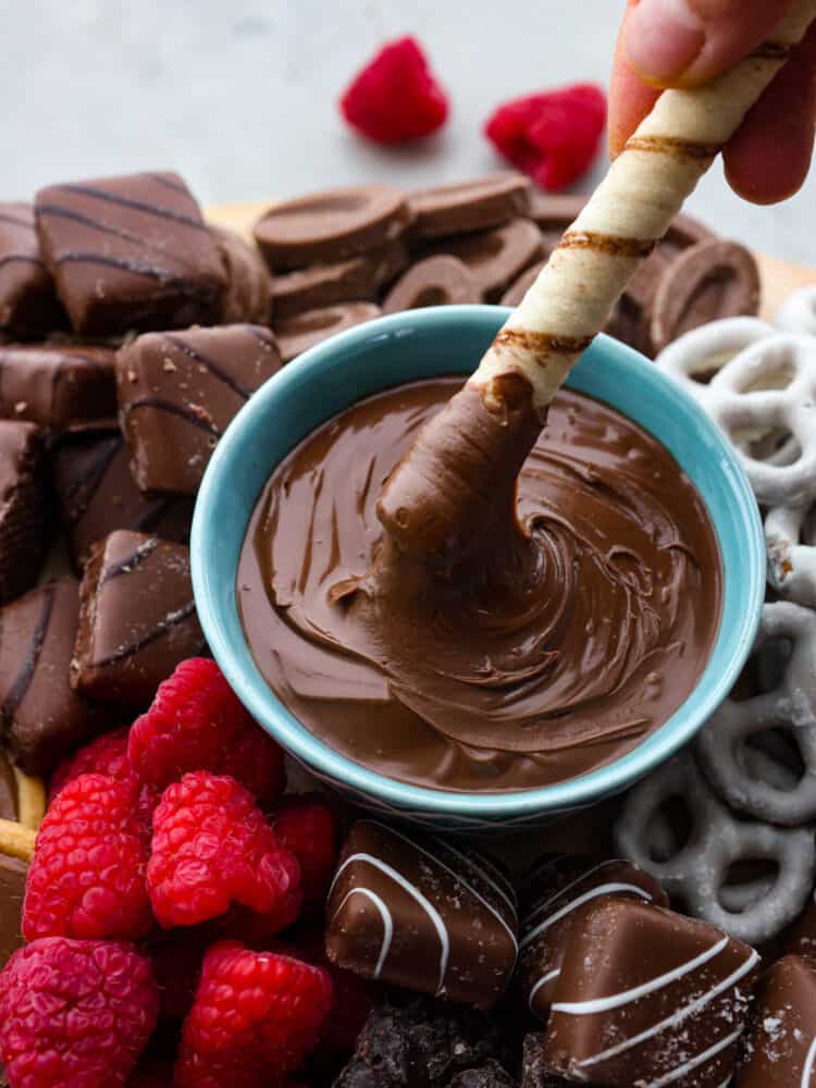 A close up photo of a cookie being dipped into a bowl of nutella. Chocolate, candy, and raspberries surround the bowl.