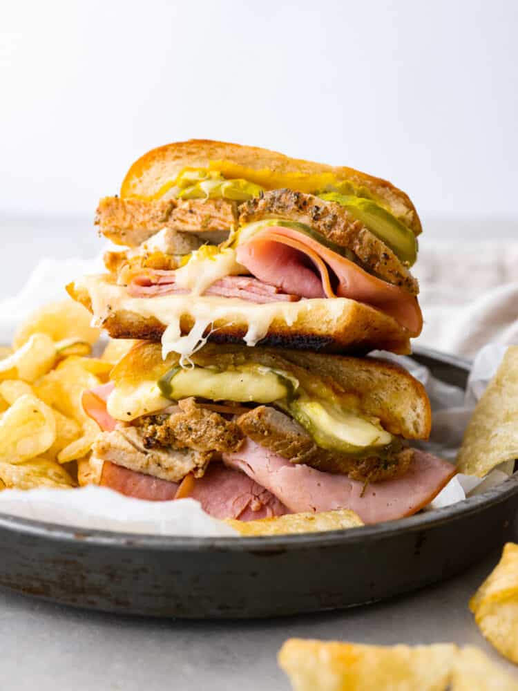 Cuban bread cut into a sandwich with lots of meat and cheese and pickles in it, with chips off to the side.