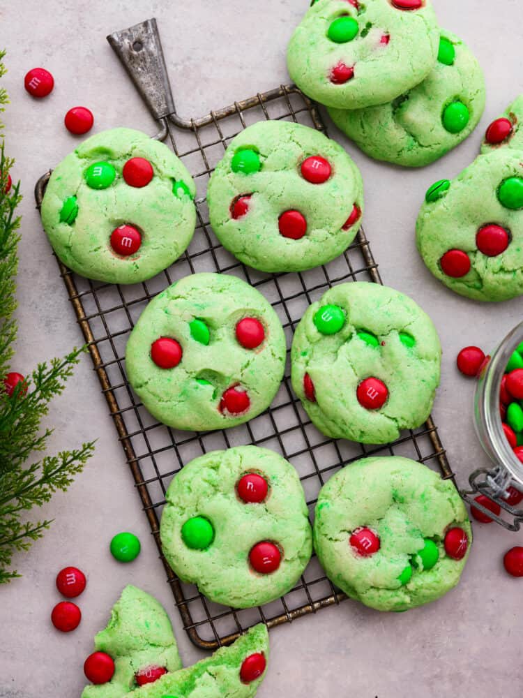 Top view of Grinch cookies on a cooling rack. Extra cookies and M&Ms are scattered around the cookies on a gray counter top.