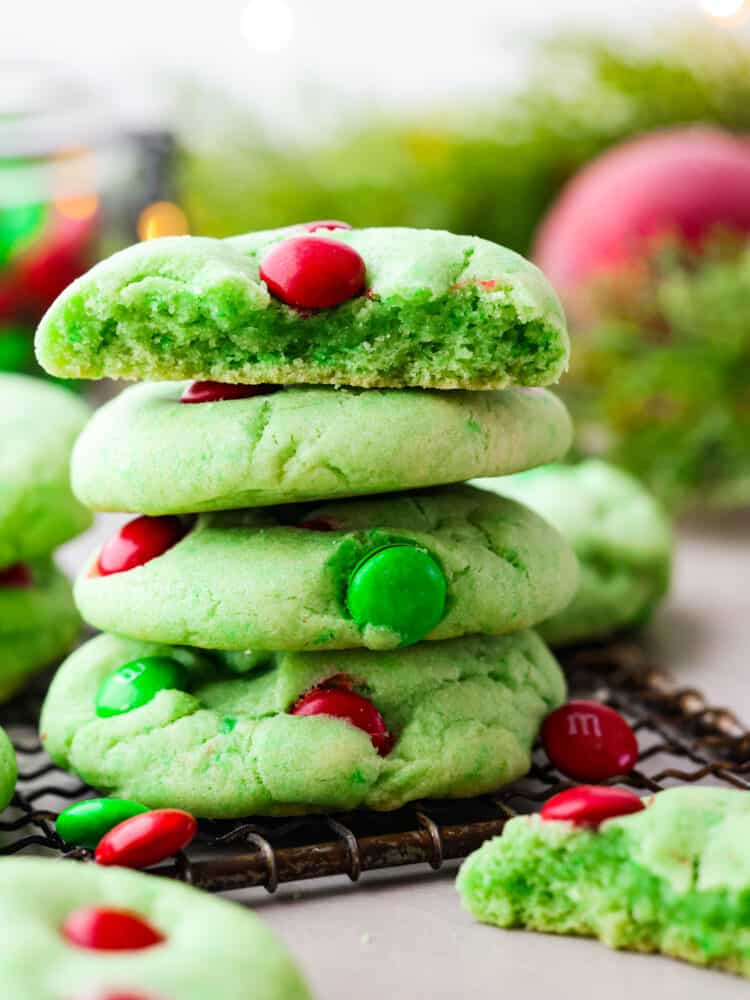 Close up side view of stacked cookies on a cooling rack. A broken cookie and Christmas ornaments are styled next to the stack of Grinch cookies.