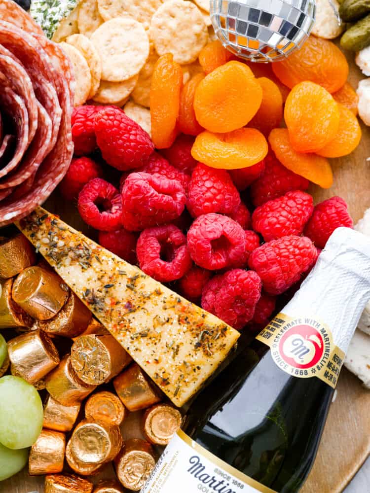 Close up view of fruit, crackers, cheese, chocolate candy, and salami rose on charcuterie board. A miniature sparkling cider and disco ball is garnished on the board.