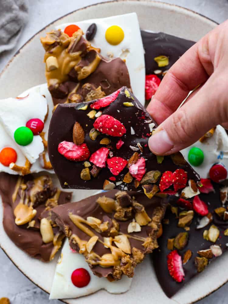 A plate of different kinds of chocolate bark with a hand grabbing a piece. 
