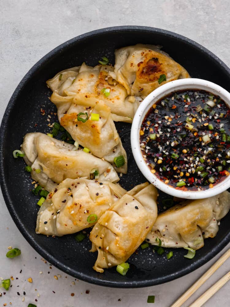 Top view of potstickers on a on a black plate with a small white bowl of sauce on the plate. Chopsticks and green onion are styled beside the plate.