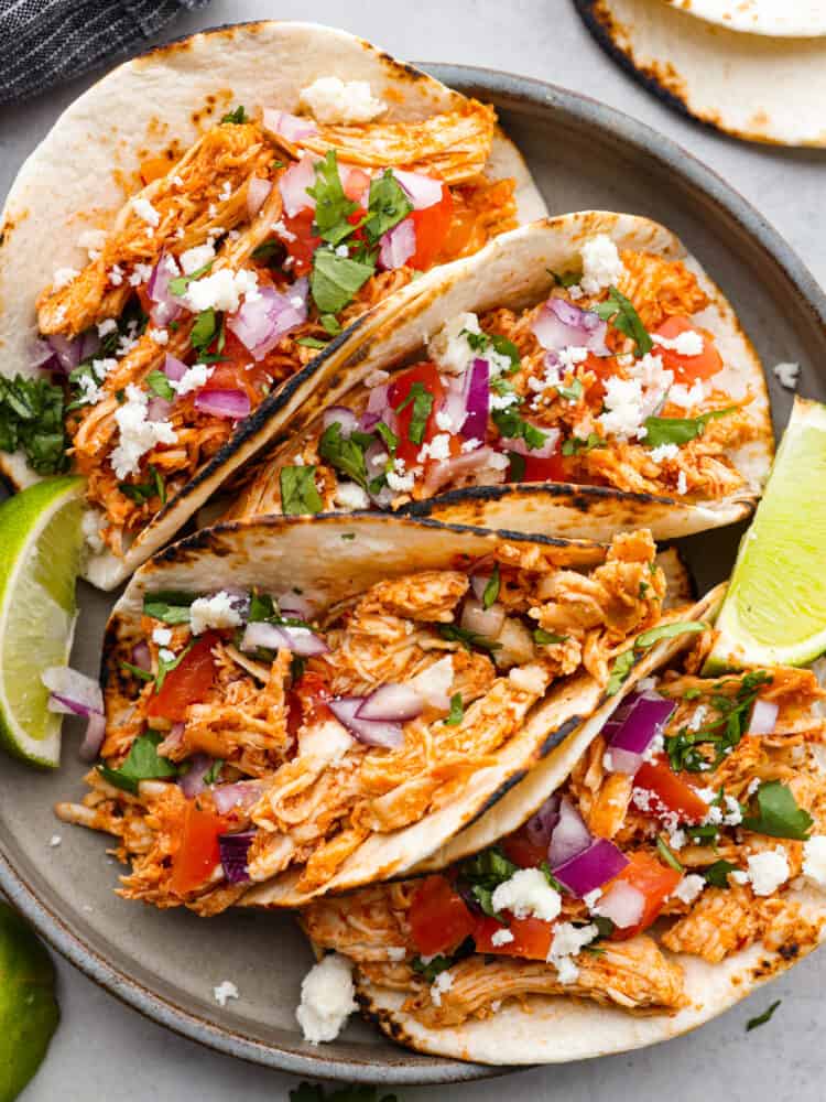 Shredded chicken tacos on a black plate with limes next to them.