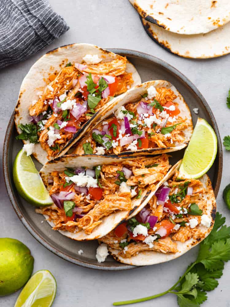 Shredded chicken tacos on a plate with limes and cilantro around it.
